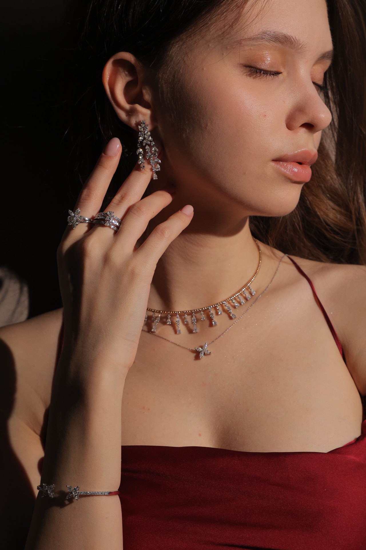 A woman in warm low light wearing layered necklaces, a drop earring, stacked rings and an open cuff bracelet